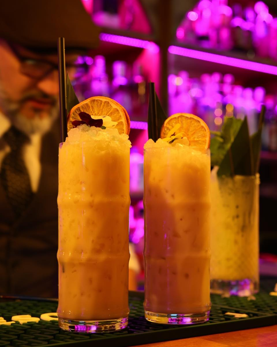 Dos cócteles tropicales con rodaja de naranja deshidratada, iluminación neón rosa de la barra