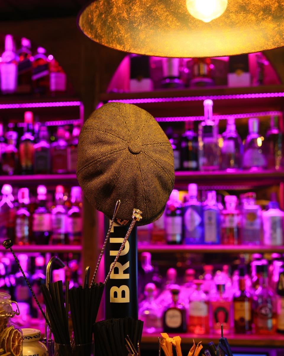 Gorra de tweed sobre un grifo de ginebra Brut, estanterías de licores con luz neón rosa
