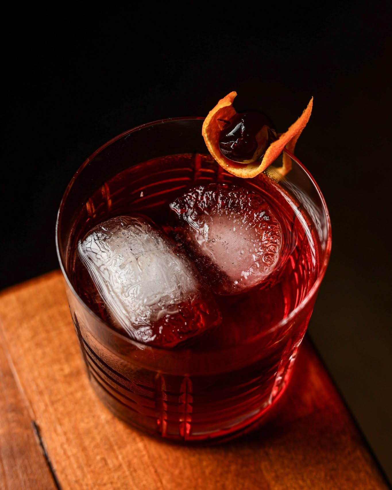 Negroni con hielo grande, guinda y piel de naranja, sobre la barra de madera
